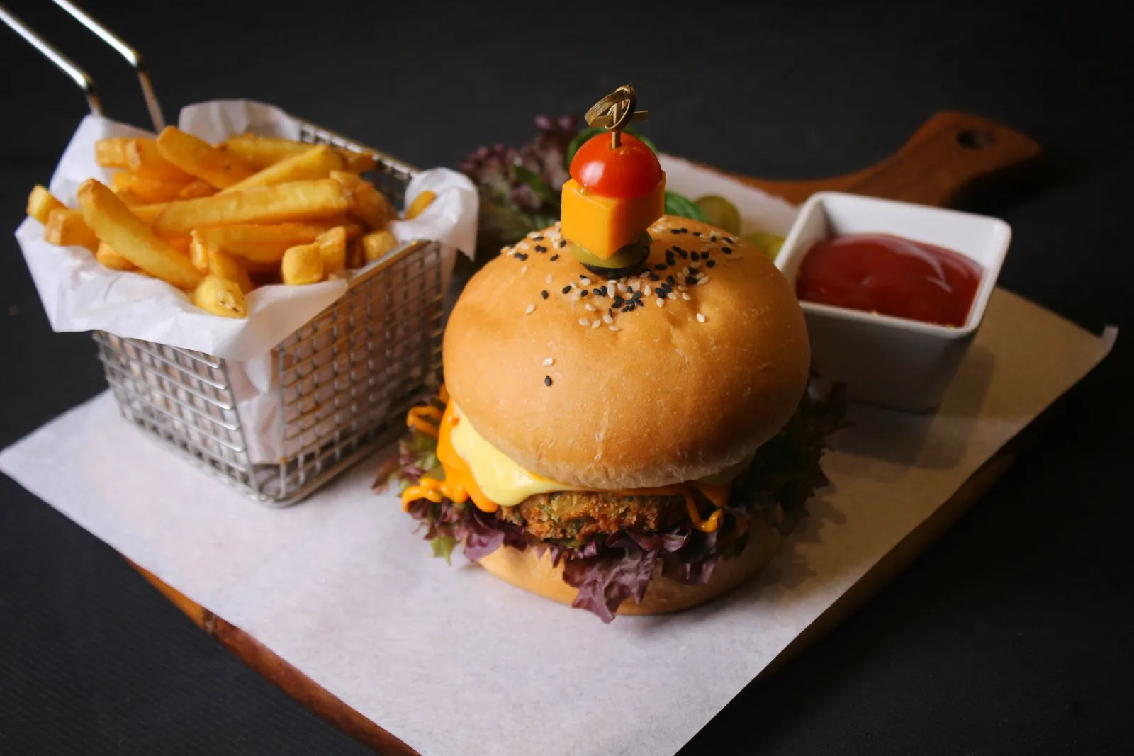 A cheeseburger with fries in a wire basket and a side of ketchup on a wooden board