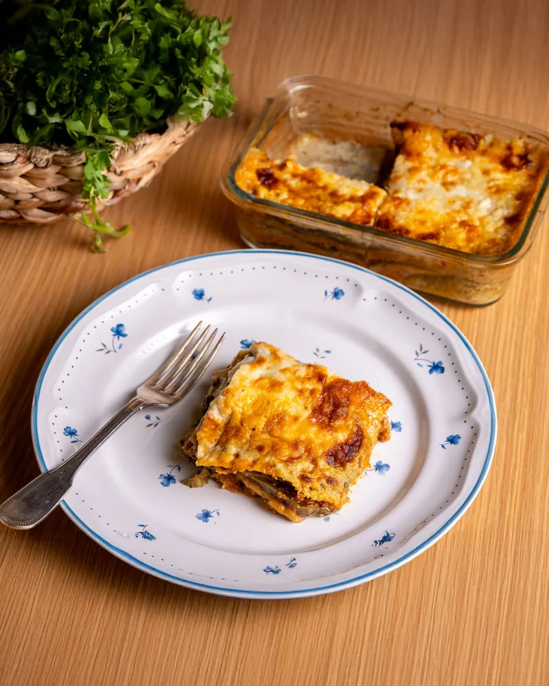A square of baked lasagna on a blue-rimmed plate, with the baking dish behind it